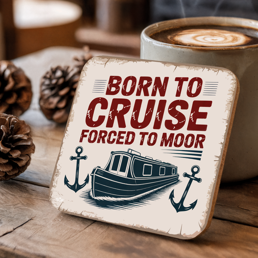 Coaster with "Born to Cruise Forced to Moor" text and boat illustration on a wooden surface with a cup of coffee.