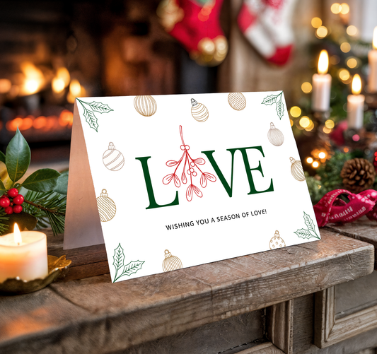 Christmas card with "LOVE" and festive decorations on a wooden surface with candles and stockings in the background.