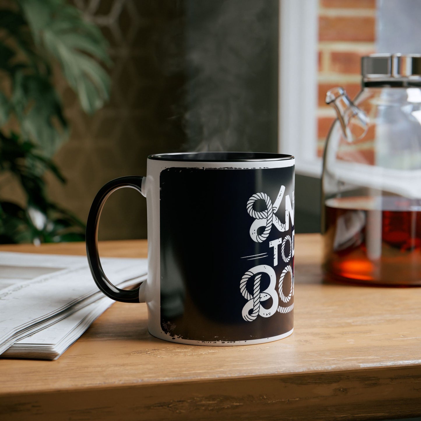 Black mug with white text on a wooden table next to a newspaper and glass container.
