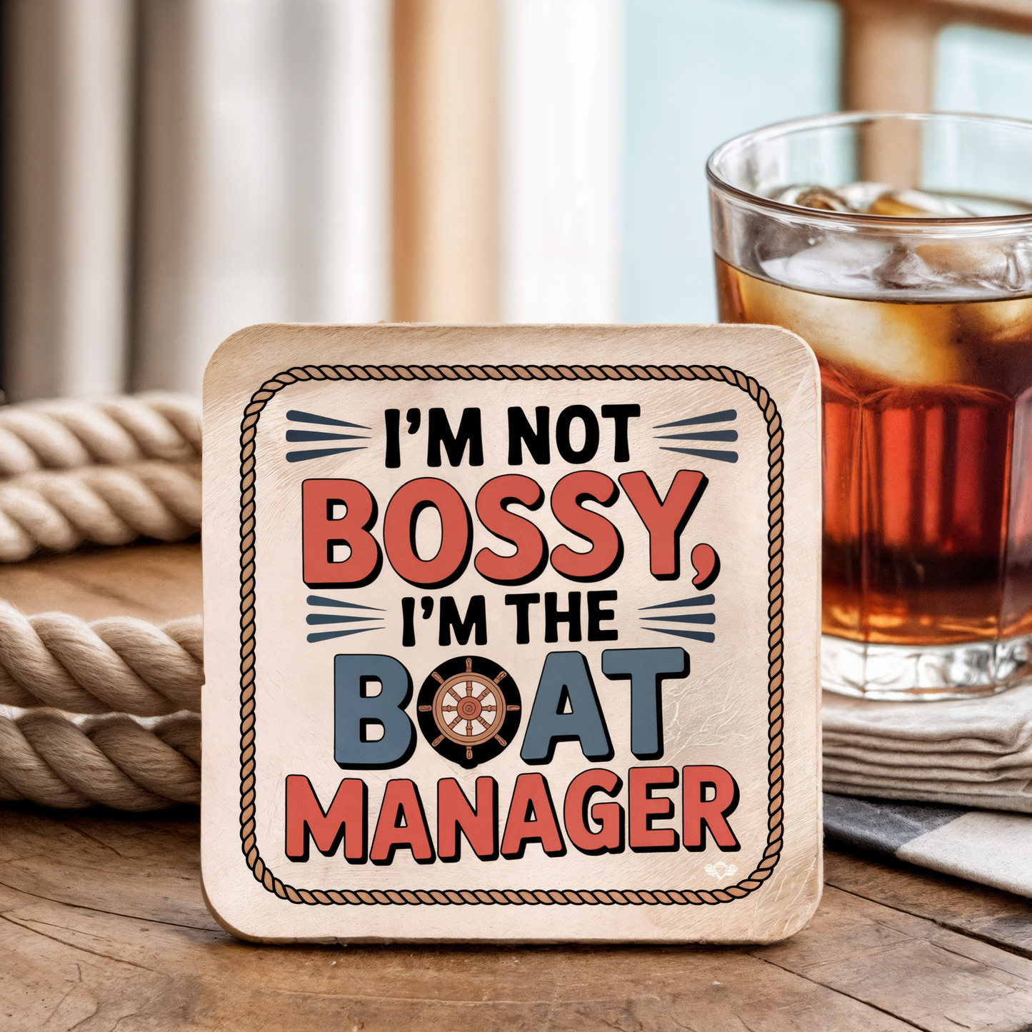 Coaster with "I'm not bossy, I'm the boat manager" text on a wooden surface with a drink in the background.