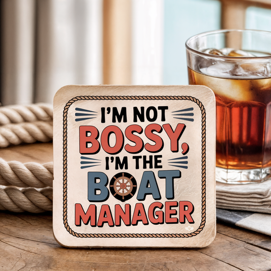 Coaster with "I'm not bossy, I'm the boat manager" text on a wooden surface with a drink in the background.